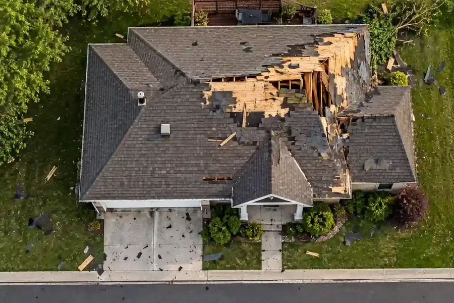 Storm damage to a shingle roof with missing shingles, exposed roof decking, and scattered debris on a residential home.